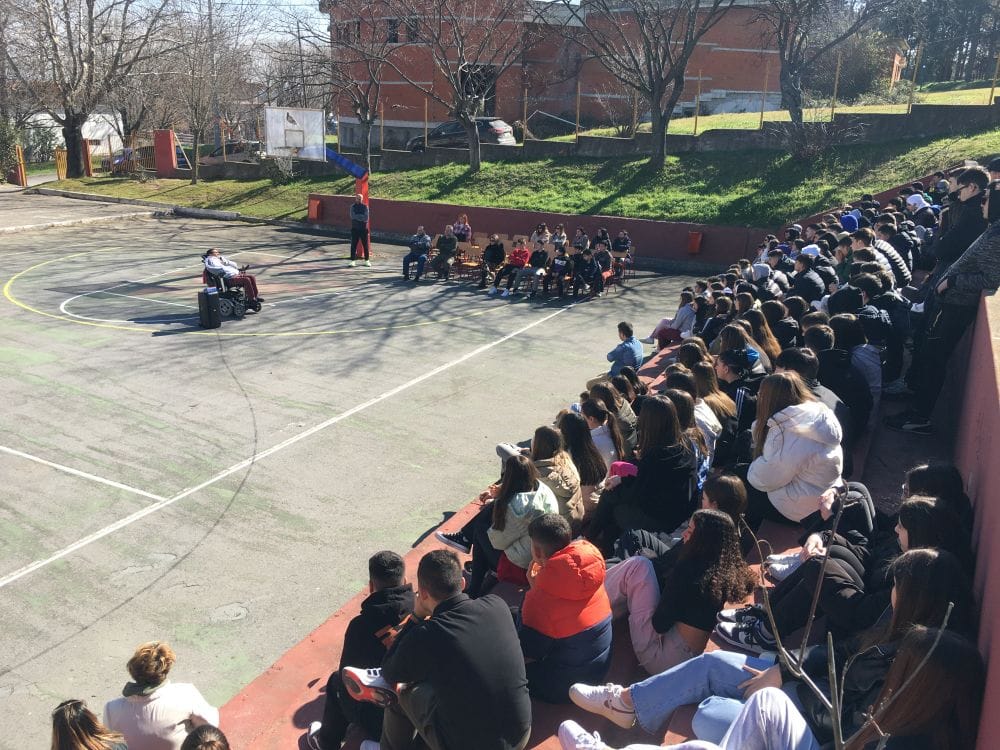 ανοιχτό γήπεδο σχολείου, μαθητές καθισμένοι στις κερκίδες, στο κέντρο ο Δημήτρης Αντωνίου
