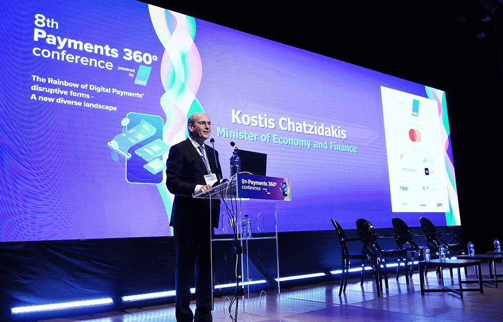 Ο υπουργός οικονομίας Κωστής Χατζηδάκις στο βήμα του συνεδρίου, στην οθόνη πίσω του το όνομά του και γραφικά σε μπλε χρώμα