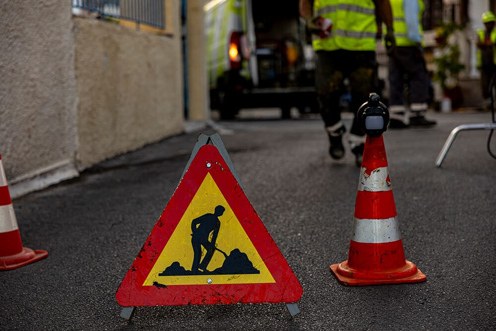 πινακίδα για έργα και κώνος ακουμπισμένα στην άσφαλτο, στο φόντο το συνεργείο του Safe Roads project