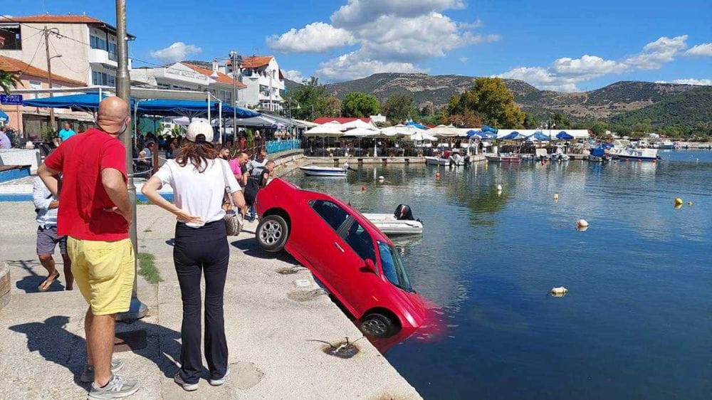 Κόκκινο αυτοκίνητο μισοπεσμένο στη θάλασσα σε λιμανάκι, γύρω γύρω κόσμος