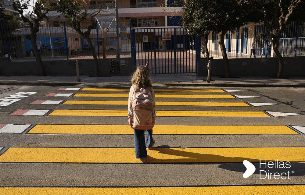 Δρόμος έξω από σχολείο, διάβαση με κίτρινα μολύβια αντί για λευκή διαγράμμιση, τη διασχίζει ένα μικρό κορίτσι που βλέπουμε πλάτη