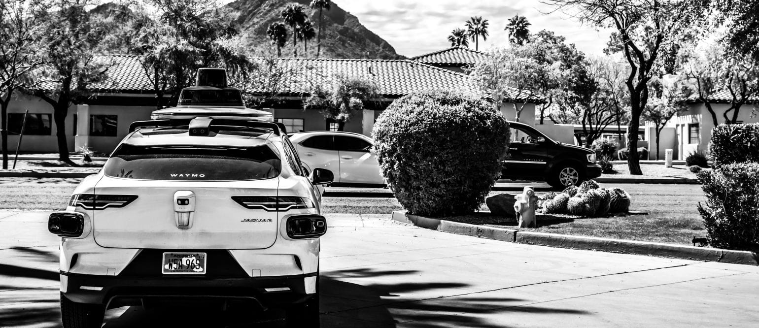 ασπρόμαυρη εικόνα, αυτοκίνητο χωρίς οδηγό από πίσω, με φόντο σπίτια και δρόμο