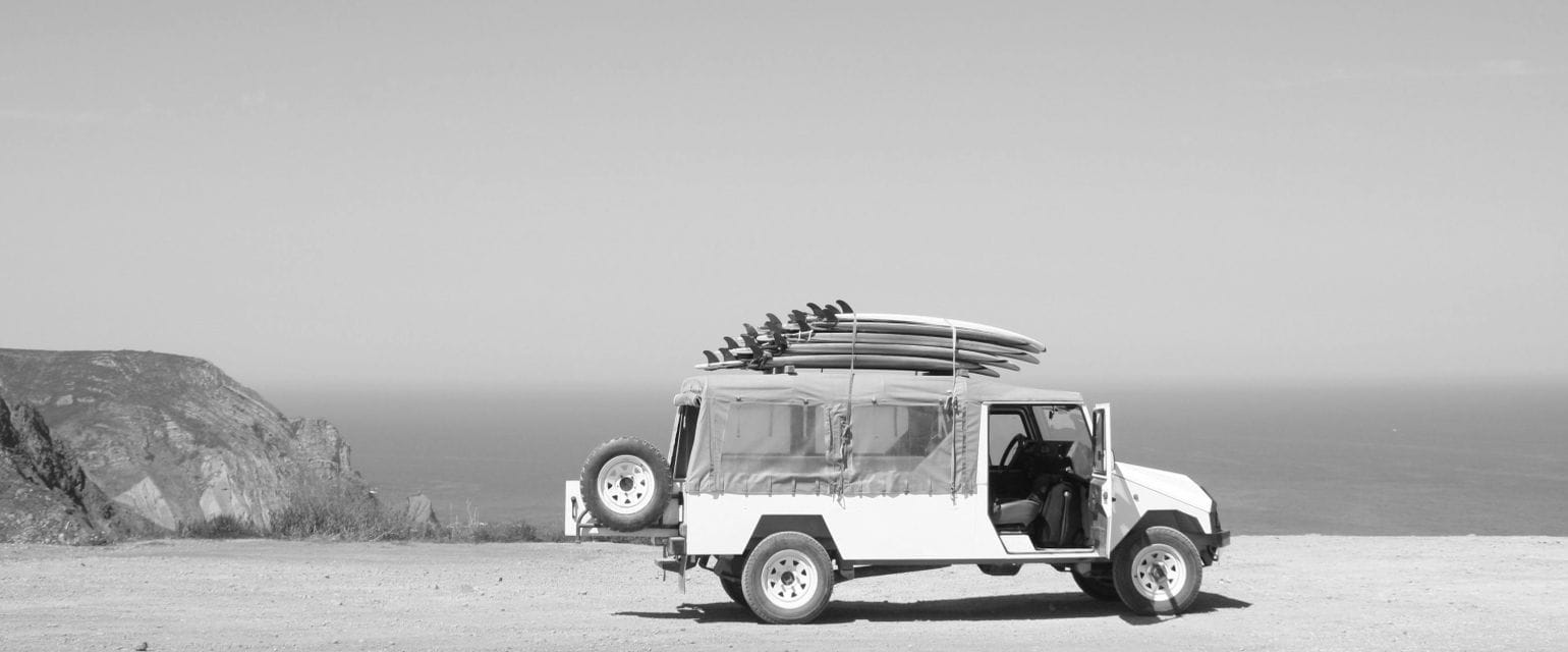 ασπρόμαυρη εικόνα, τζιπ με ανοιχτή πόρτα φορτωμένο με σανίδες σκι παρκαρισμένο με φόντο βράχια και θάλασσα